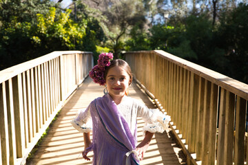 Portrait of a pretty girl dancing flamenco in a dress with frills and fringes typical of gypsies, walking on a wooden bridge in a famous park in Seville, Spain. Her hair is tied back with a flower.