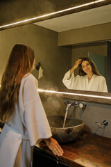 Young attractive woman standing in front of bathroom mirror, wearing white bath robe, touch the...
