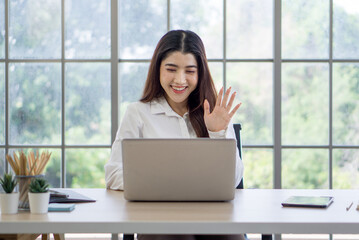 Young asian woman in white shirt sitting at a desk with laptop, smiling and waving at the screen, engaged in a video call or virtual meeting. Tablet, stationary item and indoor plant are on the table.
