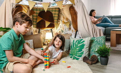 Happy little girl putting piece of stacking game while play with child and relaxed woman reading book sitting on sofa. Family having fun playing and enjoying in camping at home. Staycation concept.