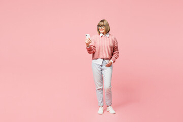Full body elderly woman 50s years old wears sweater shirt casual clothes glasses hold in hand use mobile cell phone isolated on plain pastel light pink background studio portrait. Lifestyle concept.