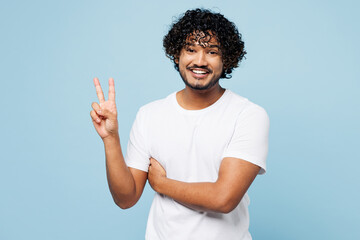 Young smiling cheerful happy Indian man he wears white t-shirt casual clothes showing victory sign look camera isolated on plain pastel light blue cyan background studio portrait. Lifestyle concept.
