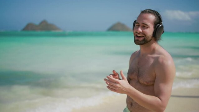 A shirtless man with headphones enjoys the rhythm, dancing with his eyes closed on a tropical beach, feeling the sun and sea breeze as he moves. Slow motion. 
