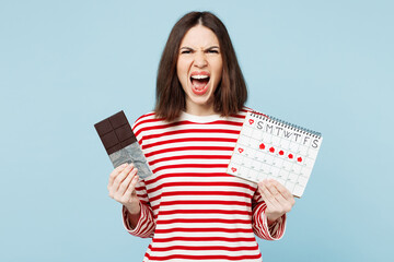 Young angry mad woman she wears red casual clothes eat sweet chocolate bar hold female periods pms calendar checking menstruation days isolated on plain blue background. Medical gynecological concept.