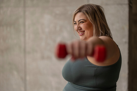 Happy curvy woman exercising with dumbbell near wall - Powered by Adobe
