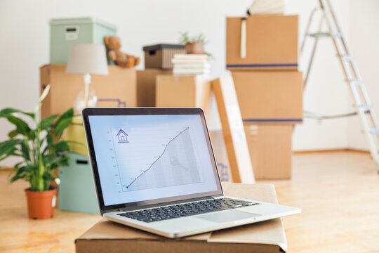 Rising line graph on laptop screen in front of cardboard boxes in an empty room in a new home
