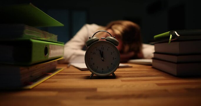 Alarm clock stands on wooden desk against sleeping lady office worker. Tired female rests at workplace feeling exhaustion from working at night