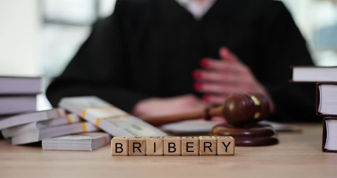 Word Bribery made of wooden cubes against stacks of money cash and judge delivering verdict. Judgement for corruption at work slow motion