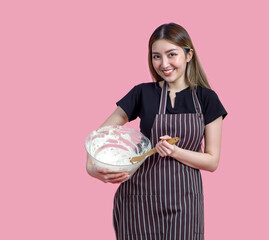 A happy woman in an apron, mixing batter in a clear bowl with a wooden spoon. Portrait on pink background with studio light. Isolated