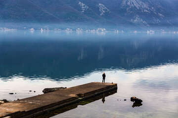 Bay of Kotor, person silhouette on pier, reflective water, serene mood for wellness ads. Bay of...