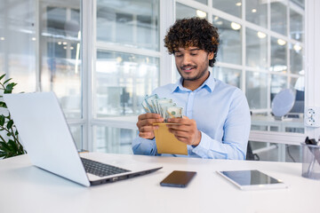 A young male professional enjoys counting a wad of cash while sitting at his sleek, contemporary...