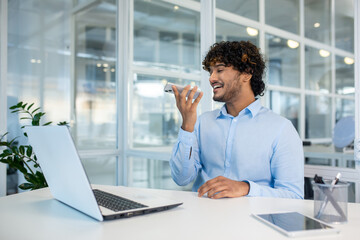 A cheerful young businessman uses his smartphone for voice commands while sitting at a desk in a bright, contemporary office setting.