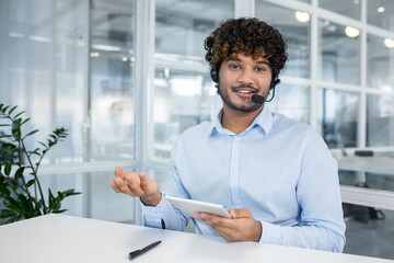 Young man in a light blue shirt and headset smiles engagingly as he talks over a video call, using a tablet. Modern office setting with glass walls.