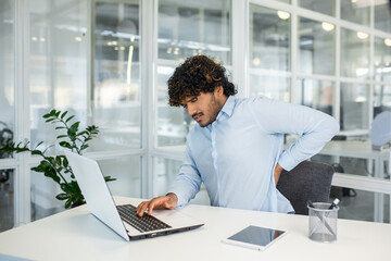 A young male professional experiences back pain while working on his laptop in a contemporary office. The image encapsulates common workplace health issues.