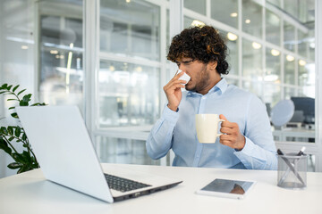 A young curly-haired man sneezing into a tissue while sipping coffee at his office desk, surrounded by modern office equipment and greenery.