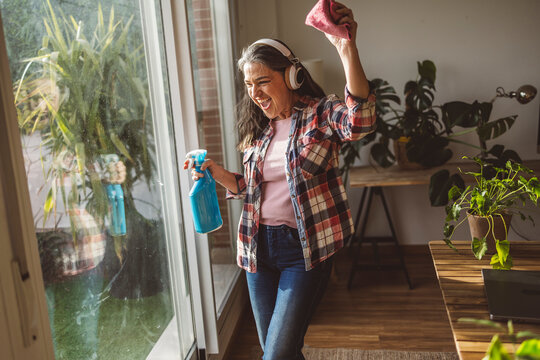Happy mature woman cleaning window glass and listening to music at home