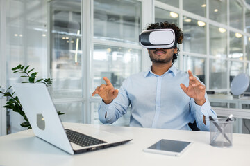 Curly-haired young professional immersed in virtual reality using a VR headset while engaging with invisible interfaces at his workplace equipped with various modern devices.