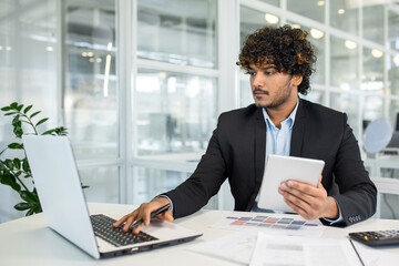A focused young man multitasks with a laptop and tablet at a desk surrounded by light-filled modern office space. He exudes professionalism and efficiency in a corporate environment.