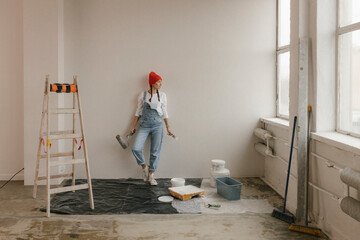 Woman leaning on white wall near ladder and paint equipment in new apartment