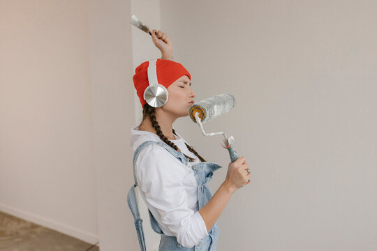 Woman listening to music through wireless headphones and singing into paint roller at new home