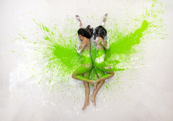Two white and green abstract painted sexy young women, doused with paint, friends, back to back, together on the floor in the artist studio