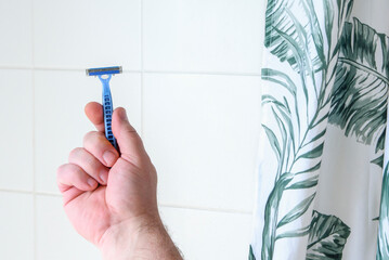 A man holding a blue razor in his hand in the bathroom against the background of a white tiled wall...