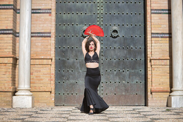 Young, beautiful, brunette woman with black top and skirt, dancing flamenco with a red fan in front of an old, black metal door. Flamenco concept, dance, art, typical Spanish.