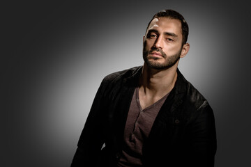Portrait of brutal brunet man in stylish jacket on dark background, Brooding young man with a serious gaze, framed by a background of dark gray tones and a rugged beard