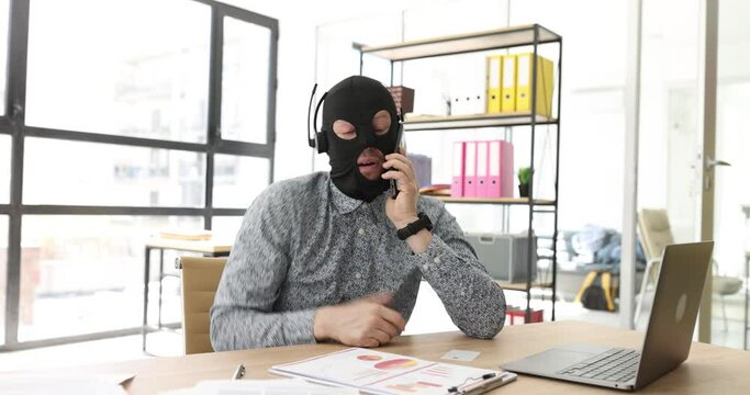 Fake manager in mask of robber talks to client sitting at workplace in company premise. Male scammer in headphones communicates cheating customer via call
