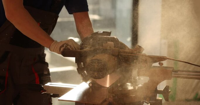 Worker uses sawing machine to cut wooden board at workplace in semi-lit room. Male specialist prepares material for parquet laying in renovated room slow motion