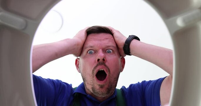 Shocked male plumber opens toilet lid putting hands on head. Man prepares to clean clogged sewer pipe. Expressive person showing surprise