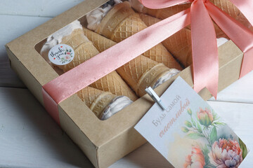 Waffel horns with marshmallows in a gift box with a transparent lid. The box is tied with a ribbon attached branded logos and a postcard.