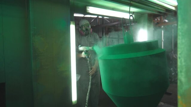 Industrial painting process in chamber at factory. Painter spraying green paint on detail in special booth. Technician in safety wear working at production. Master paint metal in specialised workshop