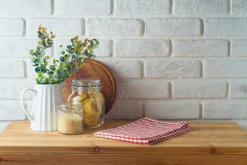 Empty wooden l table with tablecloth, plant, food jars and cutting board over white brick wall ...