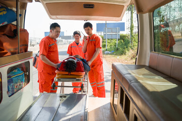 Group of paramedic or emergency medical technician (EMT) in an orange uniform places a neck and head accident victim on a bed in an ambulance. Urgent assistance during road accident.