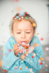 A sweet girl holds confetti in her hands and blows it away. She wears a bright smile as she enjoys the playful moment, creating an atmosphere of joy and excitement. The colorful confetti flies through