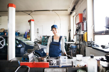 Fototapeta premium Female auto mechanic repairing, maintaining car. Beautiful woman standing in a garage, wearing blue coveralls.