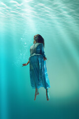 Rebirth. Tender young redhead girl in elegant dress diving into blue water, swimming in direction to sunlight. Concept of surrealism, beauty, mystery and fantasy, freedom