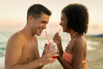 Multiethnic couple sharing a cocktail on the beach - Romantic sunset moments - Joyful interaction...