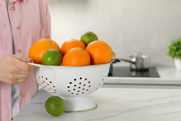 Woman holding colander with fresh fruits at white marble table in kitchen, closeup. Space for text