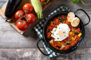 Vegetable pisto manchego with tomatoes, zucchini, peppers, onions,eggplant and egg, served in frying pan on wooden table. Top view