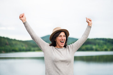 Portrait of beautiful mature woman with hat on vacation. Hands in the air, happiness.