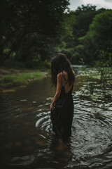 Fototapeta premium woman in a black silk dress, standing on the river bank with her back to the camera, looking down at the water and touching it with one hand, wet hair, wet skin, dark mood, natural light