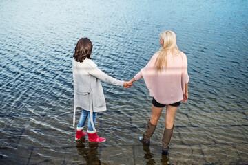 Rear view of adult daughter and mother standing in water lake reservoir, wearing rubber rain boots....
