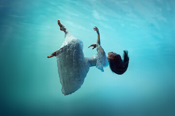 Captivating beauty surrounded by gentle waves of blue ocean. Tender girl falling down the ocean, levitating underwater. Concept of surrealism, beauty, mystery and fantasy, freedom