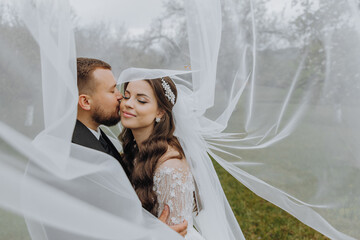a handsome and stylish groom embraces his bride in a white dress under her veil
