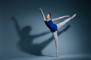 Young ballerina in bodysuit and pointe shoes performing Arabesque pose against of herself shadow on blue background.