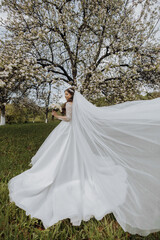 a beautiful bride in a white dress with a long veil stands near a blooming tree and holds a bouquet of flowers in her hands. Spring wedding