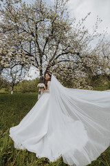 a beautiful bride in a white dress with a long veil stands near a blooming tree and holds a bouquet of flowers in her hands. Spring wedding