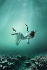 Beautiful young girl with flowing hair wears a white dress while floating above coral reefs...